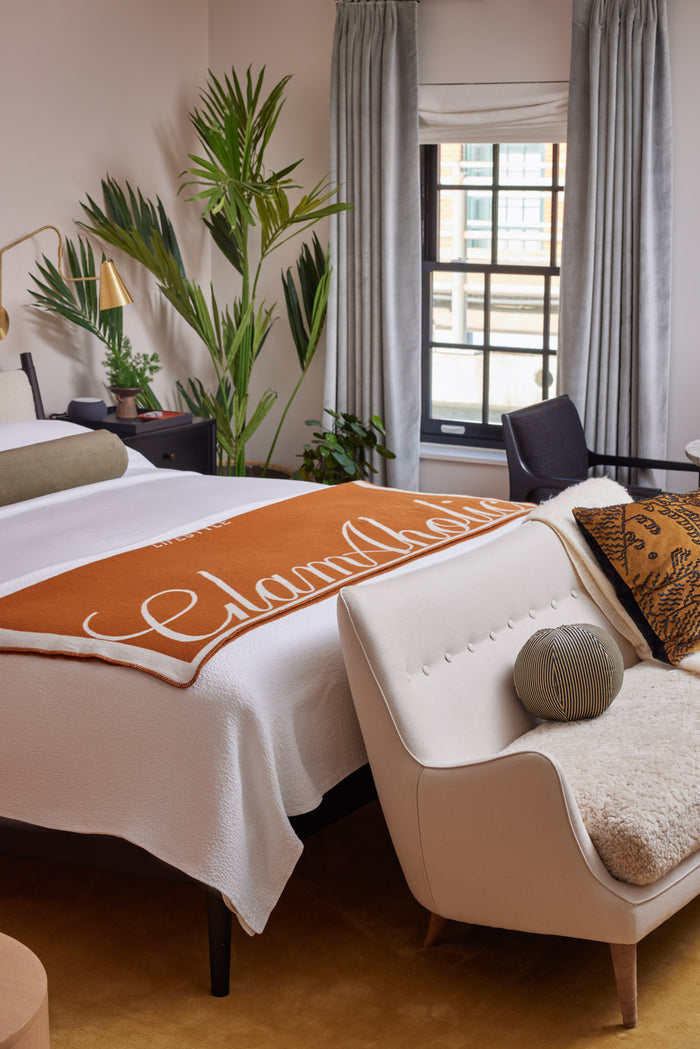 Brown luxury throw blanket with white logo draped over a white bed in a stylish, cozy bedroom setting.