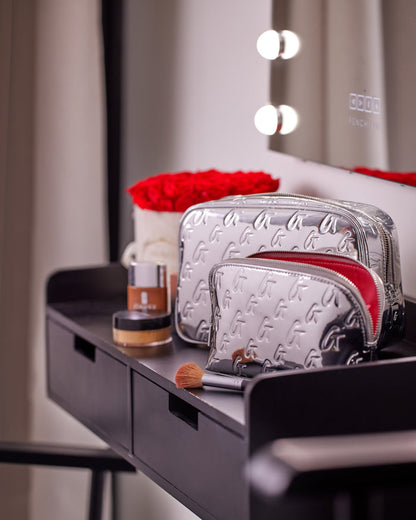 Small metallic silver monogram toiletry bag with embossed G pattern, placed on a black vanity table with makeup items.