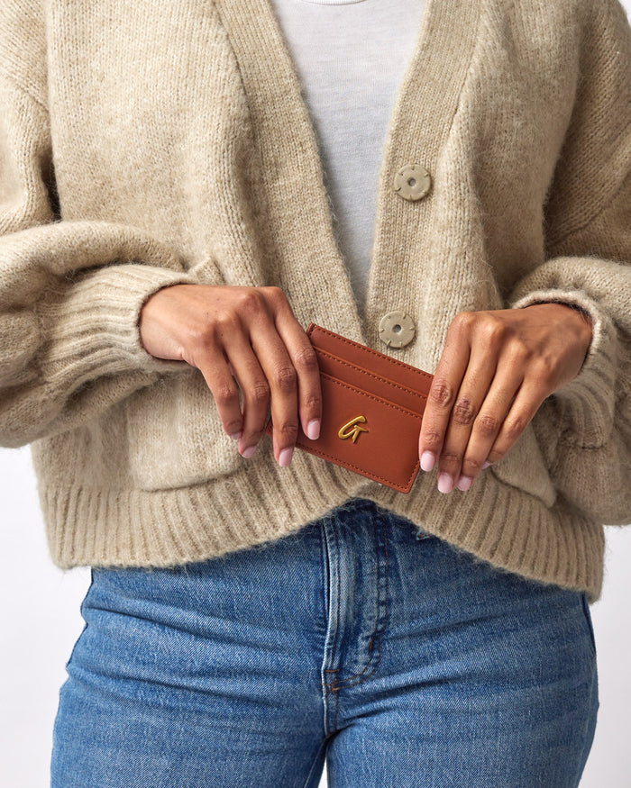 Brown leather card holder with a gold "G" logo, held by a person wearing a beige cardigan and blue jeans.