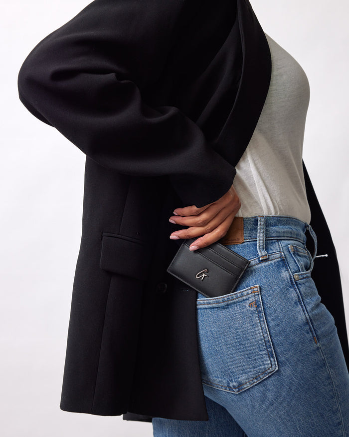 A person wearing a black coat and jeans, holding a black gunmetal card holder with a silver "G" logo near their pocket.