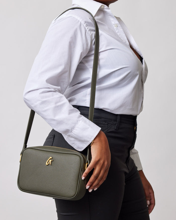 Olive green Pebble Camera Bag with a gold "G" logo, worn crossbody by a person in a white shirt and black pants.