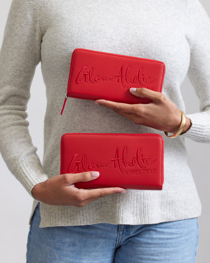 Red signature logo wallets with "Glam-Aholic" embroidery, held by a person in a beige sweater and blue jeans.