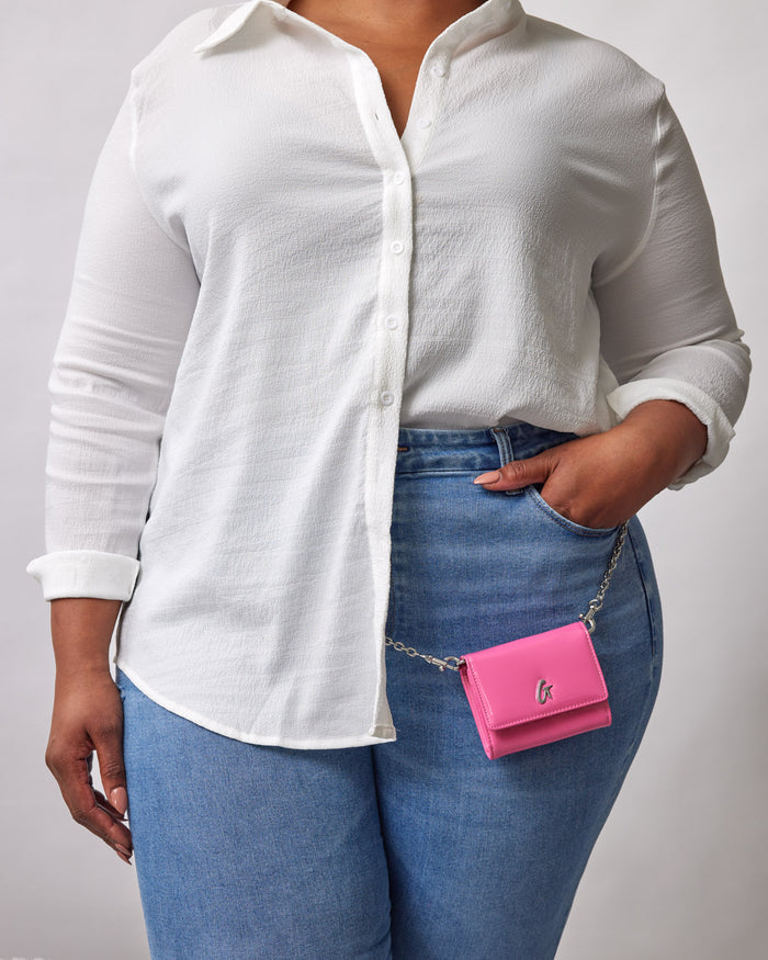 A woman wearing a white textured shirt and blue jeans, holding a small pink compact mirror with a chain, labeled "STANDARD SILVER COMPACT WOC - MIRROR HOT PINK.