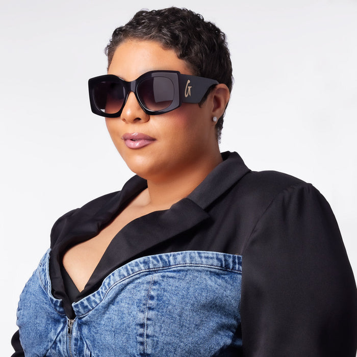 Stylish black oversized sunglasses with a gold logo, worn by a woman with short hair and a chic outfit.