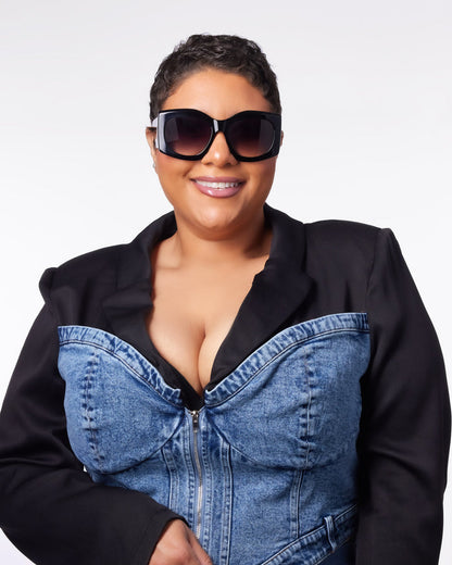 Stylish woman wearing oversized black sunglasses and a denim corset jacket, smiling confidently against a white background.
