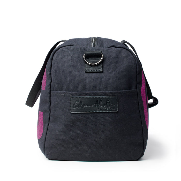 Black canvas duffle bag with pink accents and a front logo patch, featuring a zip closure and adjustable strap.
